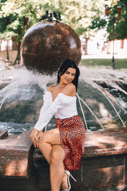A smiling and cheerful girl in stylish clothes is resting near a fountain in the city center. Lifestyle concept.  - Photo, Image