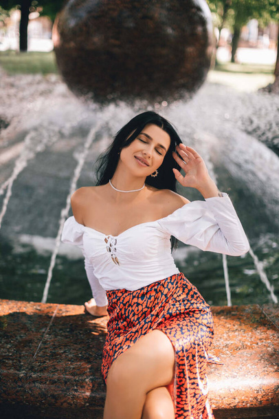 A smiling and cheerful girl in stylish clothes is resting near a fountain in the city center. Lifestyle concept.  - Foto, Bild