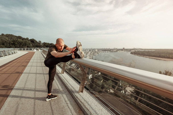 Nuori eurooppalainen mies venyttelee jalkojaan kaupungin sillalla. Terveiden elämäntapojen käsite - Valokuva, kuva