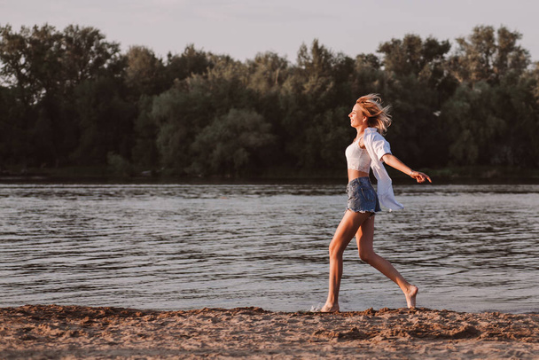 A young woman running freedom against the background of the river and trees. Profile photo a beautiful happy blonde in white clothes and denim shorts runs on the sand - Фото, изображение