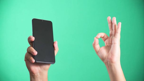 A smartphone held in the air and a hand showing the "okay" sign on a green background - Photo, image