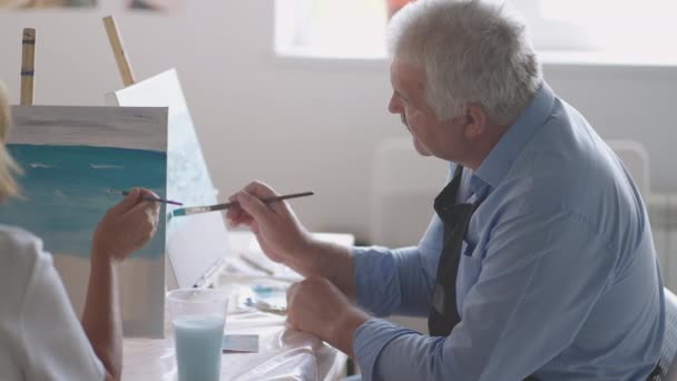 Naisopettaja näyttää eläkeläiselle, miten piirretään kuva maaleilla ja harjalla vanhusten kursseilla. Vanhempi mies piirtää kuvan eläkeläisten ryhmälle. - Materiaali, video