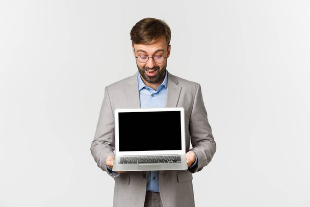Retrato de hombre de negocios barbudo en traje gris y gafas, mostrando y mirando la pantalla del ordenador portátil emocionado, demostrar gráfico, de pie sobre fondo blanco - Foto, imagen