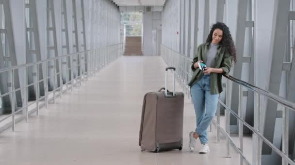 Young caucasian lady girl woman female passenger tourist traveler with suitcase stands in airport terminal holds boarding pass passport plane ticket checks time schedule on mobile phone goes to board - Footage, Video