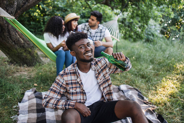 Cheerful african guy drinking beer during picnic on nature - Фото, изображение