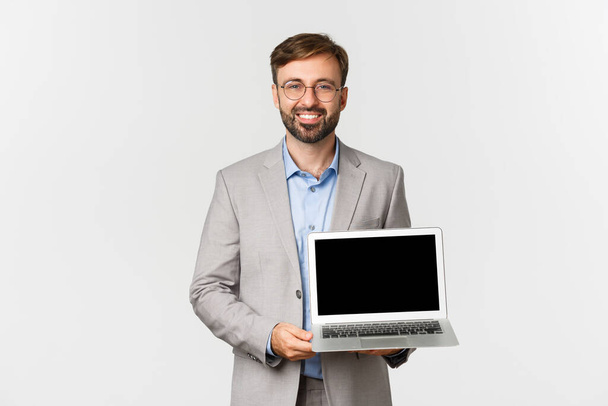 Immagine di un bellissimo uomo d'affari sorridente in abito grigio e occhiali, che mostra lo schermo del computer portatile, mostra presentazione o pubblicità, in piedi su sfondo bianco - Foto, immagini