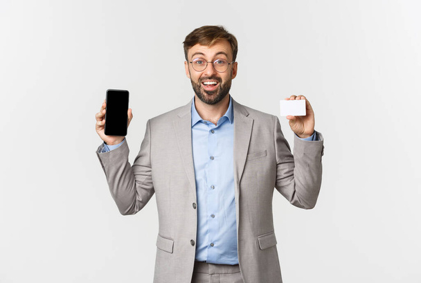 Alegre empresario masculino con barba, vestido con traje gris y gafas, mostrando aplicación en la pantalla del teléfono móvil y tarjeta de crédito, sonriendo emocionado, de pie sobre fondo blanco - Foto, Imagen