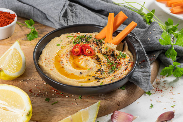 Delizioso hummus in fiocco di ceramica con bastoncino di carota immerso nell'hummus. Colorata composizione snack su sfondo bianco. - Foto, immagini