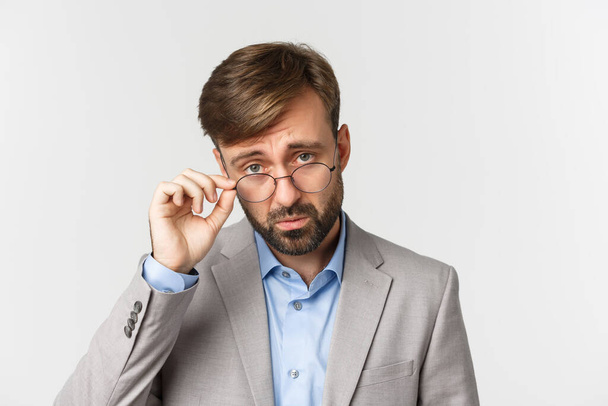 Κοντινό πλάνο του σκεπτικού γενειοφόρου άνδρα, κοιτάζοντας κάτω από τα γυαλιά και συνοφρυωμένος απογοητευμένος, στέκεται σε γκρι κοστούμι πάνω από λευκό φόντο - Φωτογραφία, εικόνα