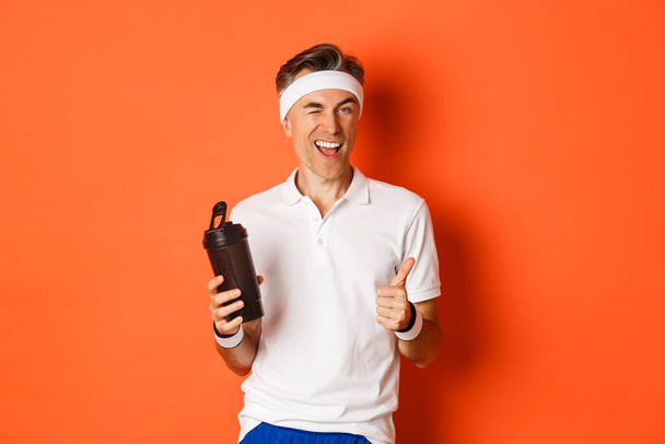 Portré magabiztos, jóképű középkorú férfi edzés egyenruhában, ivóvíz, kacsintás és mutatja hüvelykujj-up jóváhagyással, felett áll narancs háttér - Fotó, kép
