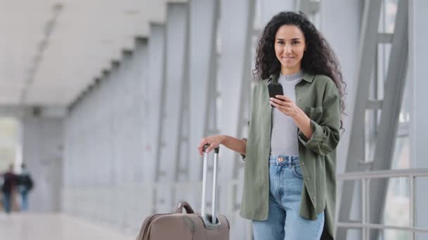 Hermosa mujer hispana usando mensajes de texto de texto de teléfonos inteligentes escribiendo mensajes de texto en el teléfono móvil verificando la hora shedule de pie en la estación de tren terminal del aeropuerto turista femenino con teléfono celular y maleta - Metraje, vídeo
