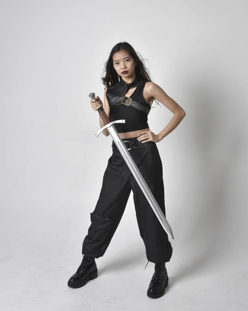 Full length portrait of pretty young asian girl wearing black tank top, utilitarian  pants and leather boots. Standing pose  holding a sword, isolated against a  studio background. - Foto, imagen