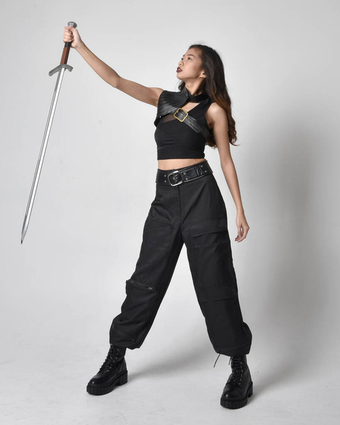 Portret młodej Azjatki w czarnej koszulce, użytkowych spodniach i skórzanych butach. Postawa stojąca z mieczem, odizolowana od tła studia. - Zdjęcie, obraz