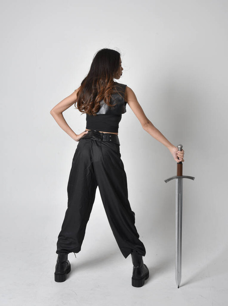 Portret młodej Azjatki w czarnej koszulce, użytkowych spodniach i skórzanych butach. Postawa stojąca z mieczem, odizolowana od tła studia. - Zdjęcie, obraz