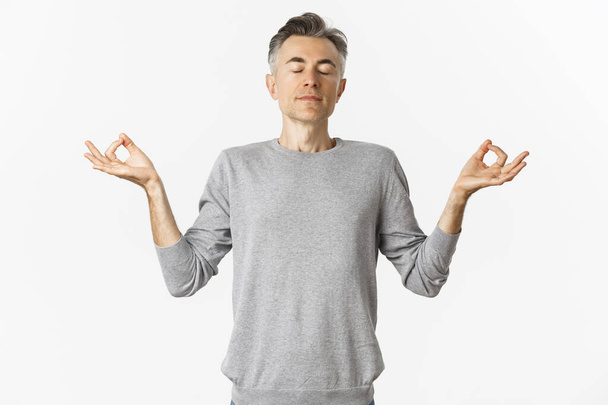 Portrait of calm and relaxed middle-aged man, breathing air freely, meditating with eyes closed and hands spread sideways, practice yoga over white background - Photo, Image