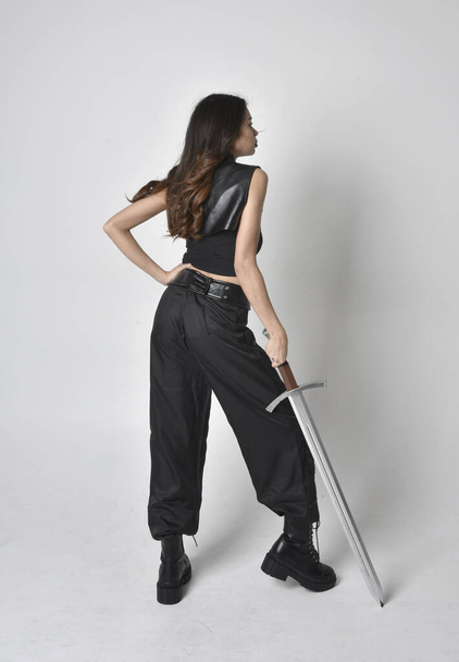 Full length portrait of pretty young asian girl wearing black tank top, utilitarian  pants and leather boots. Standing pose  holding a sword, isolated against a  studio background. - Fotografie, Obrázek