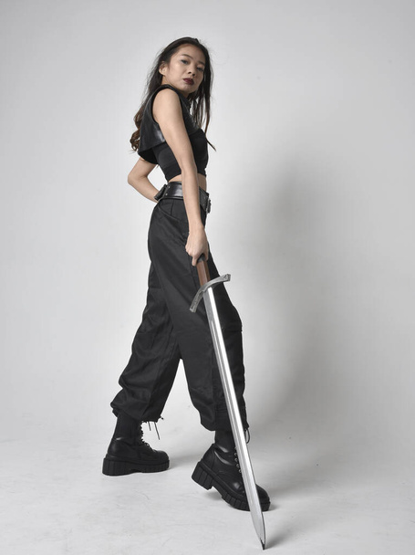 Full length portrait of pretty young asian girl wearing black tank top, utilitarian  pants and leather boots. Standing pose  holding a sword, isolated against a  studio background. - 写真・画像