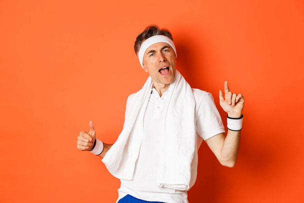 Image d'un homme de fitness d'âge moyen heureux et insouciant, dansant après l'entraînement, tenant une serviette au-dessus du cou, posant sur un fond orange - Photo, image