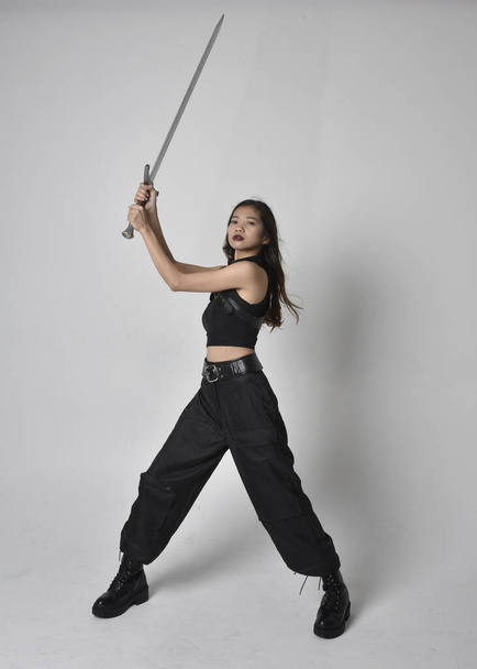 Full length portrait of pretty young asian girl wearing black tank top, utilitarian  pants and leather boots. Standing pose  holding a sword, isolated against a  studio background. - Фото, зображення