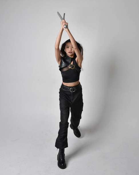 Full length portrait of pretty young asian girl wearing black tank top, utilitarian  pants and leather boots. Standing pose  holding a sword, isolated against a  studio background. - Zdjęcie, obraz