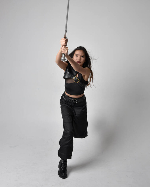 Full length portrait of pretty young asian girl wearing black tank top, utilitarian  pants and leather boots. Standing pose  holding a sword, isolated against a  studio background. - Valokuva, kuva
