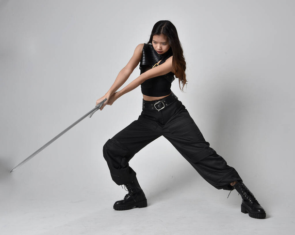 Full length portrait of pretty young asian girl wearing black tank top, utilitarian  pants and leather boots. Standing pose  holding a sword, isolated against a  studio background. - Fotó, kép