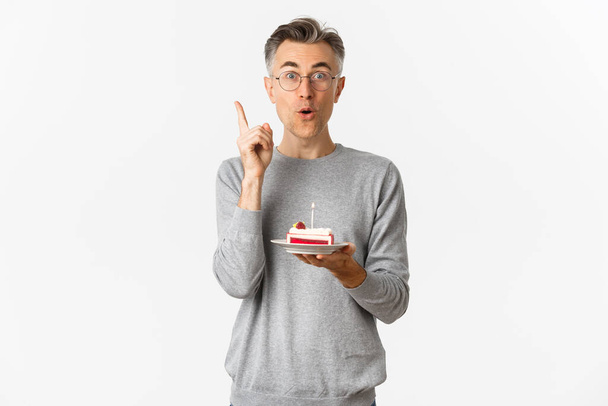 Immagine di felice uomo di mezza età, che celebra il compleanno, hanno grande idea di cosa desiderare mentre soffia candela sulla torta b-day, in piedi su sfondo bianco - Foto, immagini