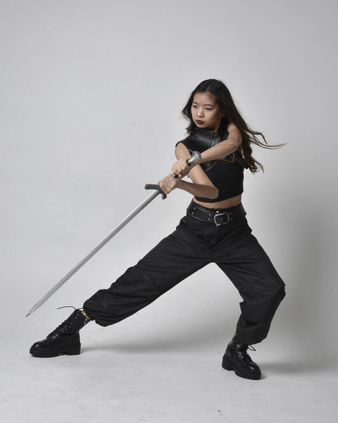 Full length portrait of pretty young asian girl wearing black tank top, utilitarian  pants and leather boots. Standing pose  holding a sword, isolated against a  studio background. - Foto, Imagem