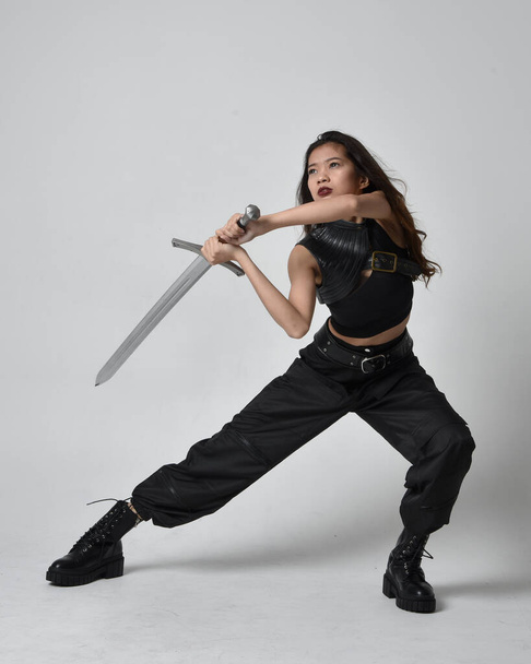 Full length portrait of pretty young asian girl wearing black tank top, utilitarian  pants and leather boots. Standing pose  holding a sword, isolated against a  studio background. - Фото, изображение