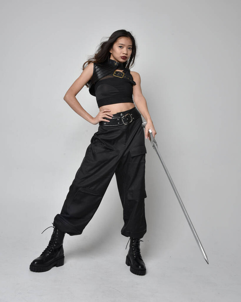Full length portrait of pretty young asian girl wearing black tank top, utilitarian  pants and leather boots. Standing pose  holding a sword, isolated against a  studio background. - Fotó, kép