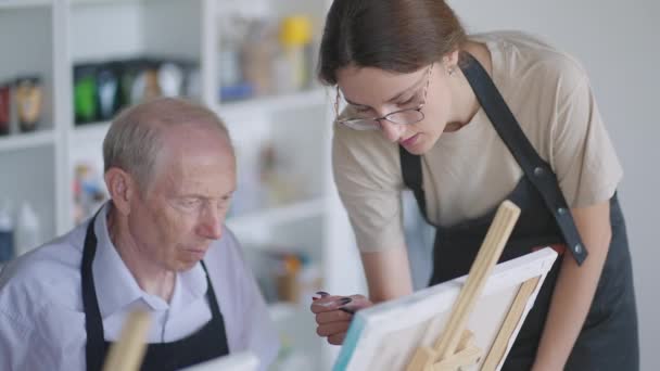 Seitenansicht einer fröhlichen Seniorin, die beim Zeichnen als Freizeitaktivität oder Therapie im Malkurs mit der Gruppe pensionierter Frauen und Männer lächelt - Filmmaterial, Video