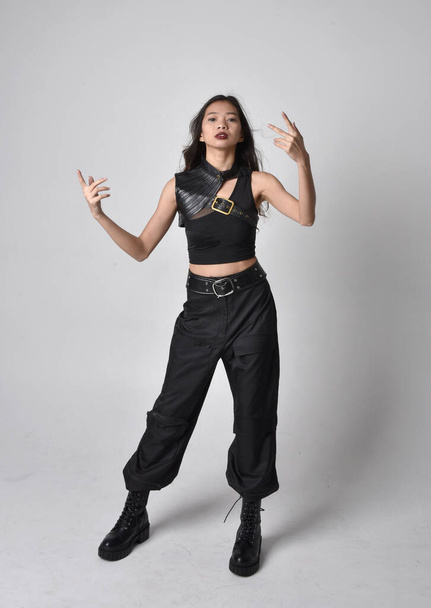 Full length portrait of pretty young asian girl wearing black tank top, utilitarian  pants and leather boots. Standing pose with gestural hands, isolated against a  studio background. - Foto, imagen