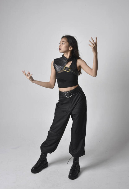 Portret młodej Azjatki w czarnej koszulce, użytkowych spodniach i skórzanych butach. Pozycja stojąca z gestykulacyjnymi dłońmi, odizolowana na tle studia. - Zdjęcie, obraz