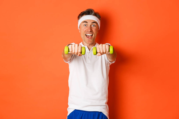 Изображение активного спортсмена средних лет, занимающегося спортивными упражнениями с гантелями и улыбающегося взволнованного, стоящего на оранжевом фоне - Фото, изображение