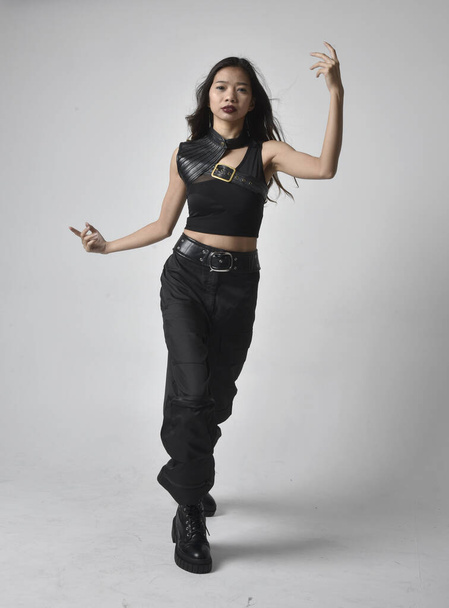 Full length portrait of pretty young asian girl wearing black tank top, utilitarian  pants and leather boots. Standing pose with gestural hands, isolated against a  studio background. - Foto, Bild