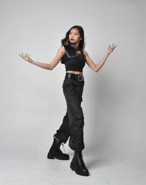 Full length portrait of pretty young asian girl wearing black tank top, utilitarian  pants and leather boots. Standing pose with gestural hands, isolated against a  studio background. - Photo, Image