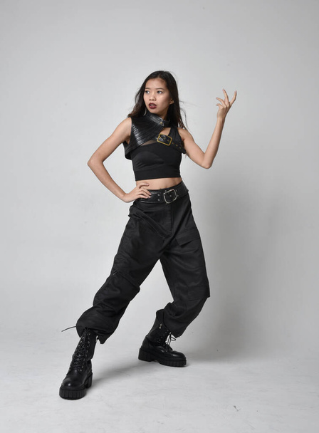 Portret młodej Azjatki w czarnej koszulce, użytkowych spodniach i skórzanych butach. Pozycja stojąca z gestykulacyjnymi dłońmi, odizolowana na tle studia. - Zdjęcie, obraz