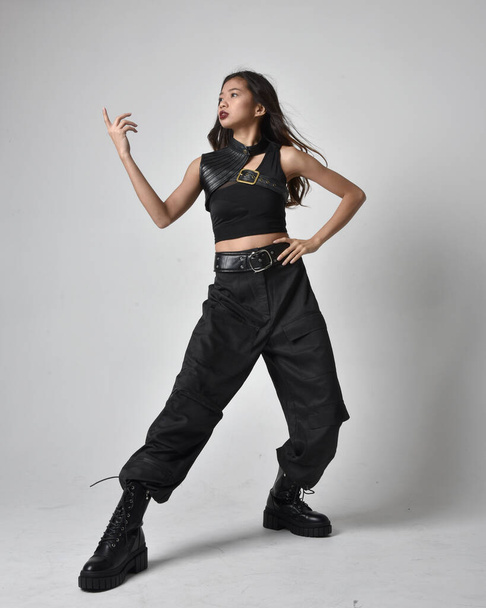 Full length portrait of pretty young asian girl wearing black tank top, utilitarian  pants and leather boots. Standing pose with gestural hands, isolated against a  studio background. - Zdjęcie, obraz