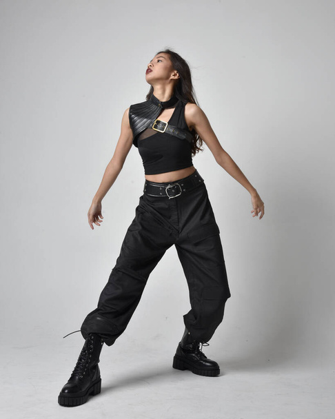 Full length portrait of pretty young asian girl wearing black tank top, utilitarian  pants and leather boots. Standing pose with gestural hands, isolated against a  studio background. - Valokuva, kuva