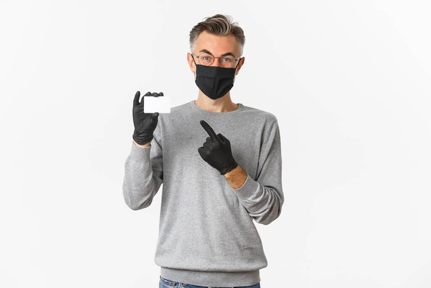 Concept of covid-19, social distancing and lifestyle. Handsome middle-aged man in medical mask, gloves and glasses, pointing finger at credit card, standing over white background - Photo, Image