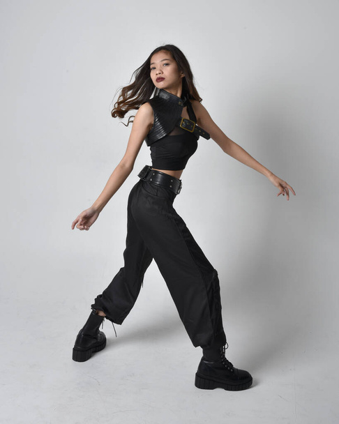 Full length portret van mooie jonge aziatische meisje dragen zwarte tank top, utilitaire broek en lederen laarzen. Staande pose met gebaren handen, geïsoleerd tegen een studio achtergrond. - Foto, afbeelding