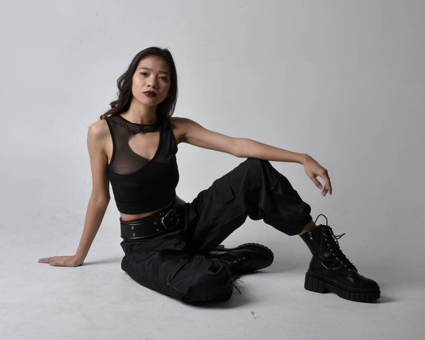 Full length portrait of pretty young asian girl wearing black tank top, utilitarian  pants and leather boots. Sitting poseisolated against a  studio background. - Фото, изображение