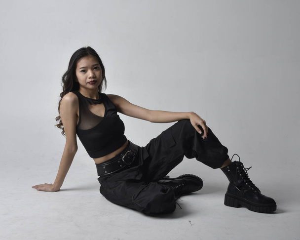 Full length portrait of pretty young asian girl wearing black tank top, utilitarian  pants and leather boots. Sitting poseisolated against a  studio background. - 写真・画像