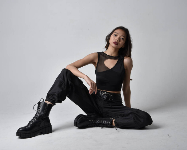 Full length portrait of pretty young asian girl wearing black tank top, utilitarian  pants and leather boots. Sitting poseisolated against a  studio background. - Foto, Imagen