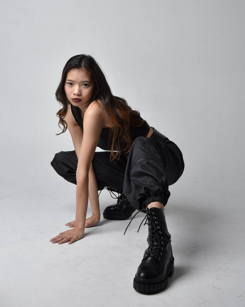 Portret młodej Azjatki w czarnej koszulce, użytkowych spodniach i skórzanych butach. Siedzenie w izolatce na tle studia. - Zdjęcie, obraz