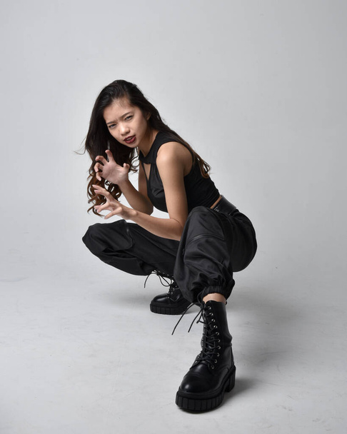 Full length portrait of pretty young asian girl wearing black tank top, utilitarian  pants and leather boots. Sitting poseisolated against a  studio background. - Foto, Imagem