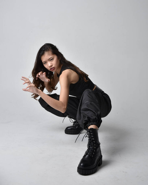 Full length portrait of pretty young asian girl wearing black tank top, utilitarian  pants and leather boots. Sitting poseisolated against a  studio background. - Foto, afbeelding