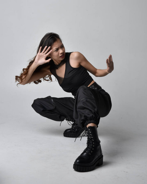 Full length portrait of pretty young asian girl wearing black tank top, utilitarian  pants and leather boots. Sitting poseisolated against a  studio background. - Zdjęcie, obraz