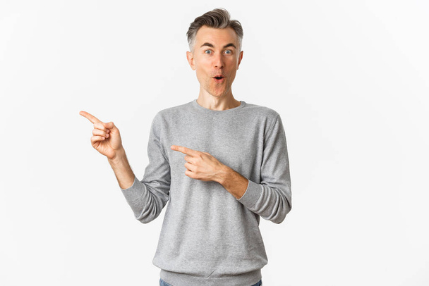 Amazed middle-aged guy in grey sweater, pointing fingers left and smiling excited, showing awesome promo banner, standing over white background - Photo, Image