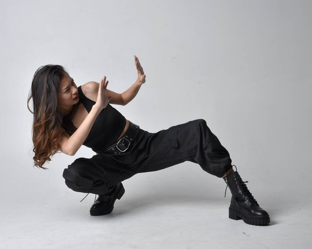 Full length portrait of pretty young asian girl wearing black tank top, utilitarian  pants and leather boots. Sitting poseisolated against a  studio background. - Foto, imagen